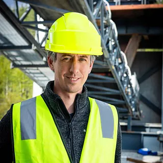 En arbeider med gul hjelm og refleksvest står foran industrielt utstyr på et pukkverk.