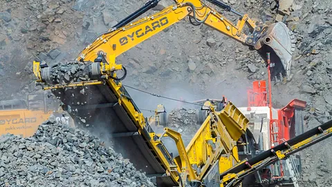 En gravemaskin laster stein på et transportbånd i et aktivt pukkverk omgitt av bratte fjellvegger.