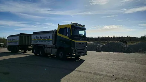 En lastebil med henger fra John Myrvang AS står på et grusområde med hauger av pukk under en solrik himmel.