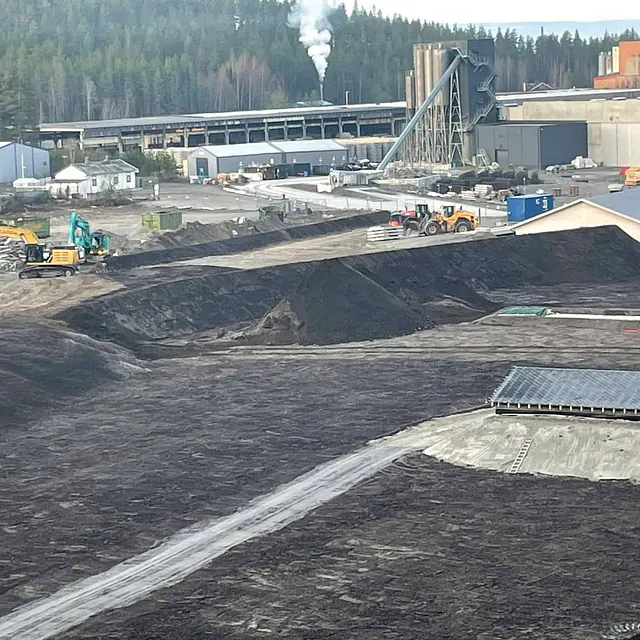 Et industriområde med gravemaskiner, jordvoller og fabrikkanlegg omgitt av skog.