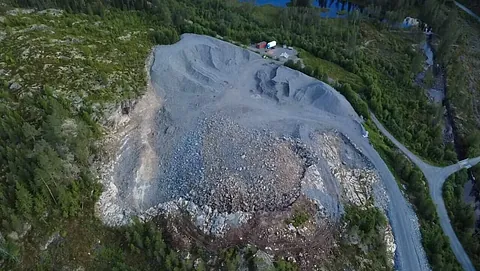 Et stort steinuttak på toppen av en høyde omgitt av skog og en grusvei.