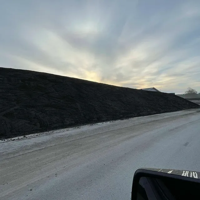 En lang voll av mørk masse langs en bred grusvei i et industriområde ved soloppgang.