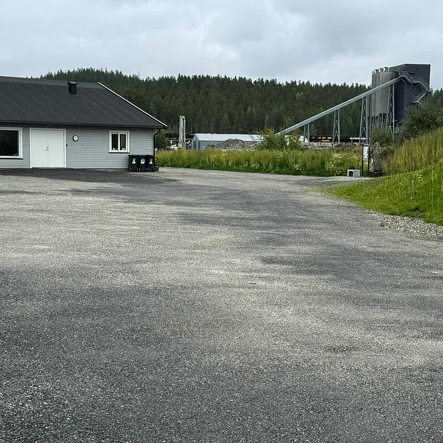 En stor grusplass foran en lav industribygning med skog i bakgrunnen og et steinprosesseringsanlegg til høyre.