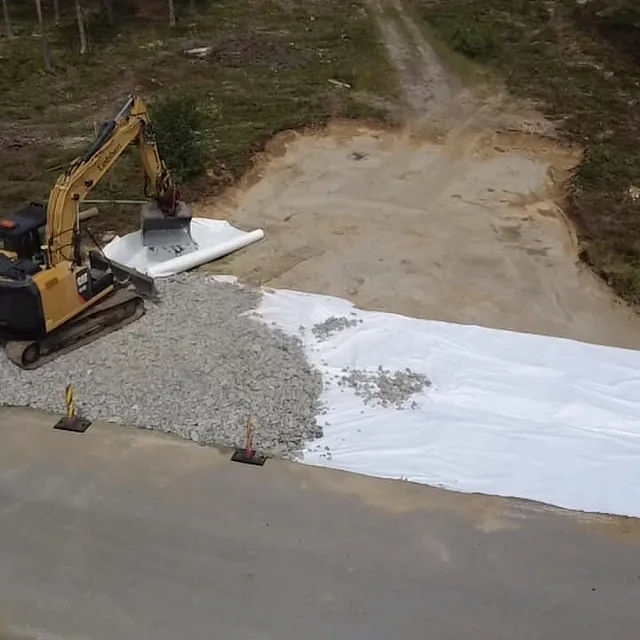 En gravemaskin legger pukk over fiberduk ved siden av en landevei i et skogsområde.
