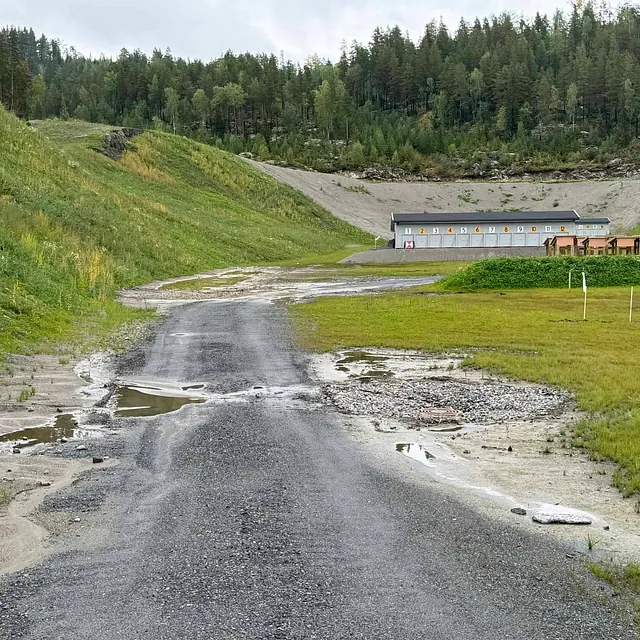 En grusvei med vanndammer leder mot en skytebane omgitt av gressbakker og skog.