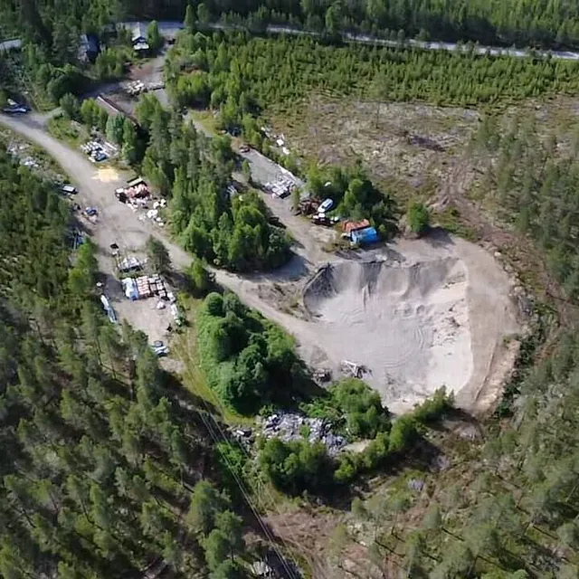 Et område i skogen med grusuttak, små bygninger og lagrede masser sett fra luften.